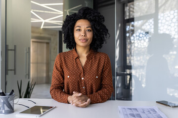 A confident young African American woman in a stylish polka dot shirt poses at her workspace with a tablet and blueprint ,in a well-lit modern office setting. - Powered by Adobe