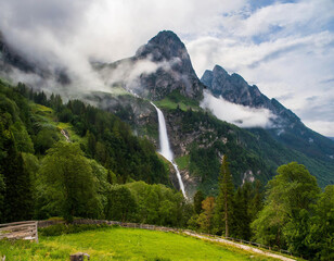 Obraz premium A mountain shrouded in mist and clouds, with a waterfall cascading from its peak, occupies the center of the photo