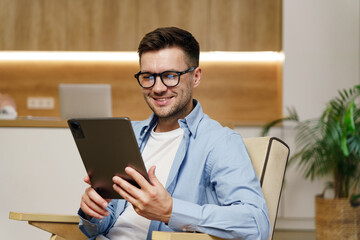 A smart man in casual attire enjoys using a tablet in a comfortable office environment with...