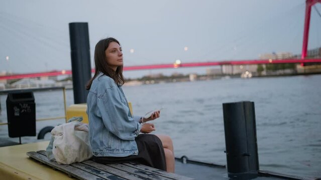young woman sitting on quay looking at lights of the evening city, admiring  view. Girl using mobile phone against  background of big city. Female  wait for water taxi or ferry
