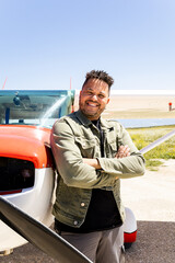 Fototapeta premium Vertical photo of a dark-skinned man poses next to a red and white plane. The Dominican adult poses with his arms crossed looking at the camera. Light aircraft concept.