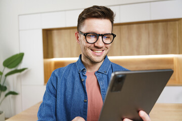 A man with glasses and a genuine smile looks at his tablet, standing in a modern kitchen with warm...
