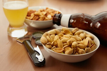 Pistachios, almonds, and cashews in bowls next to a bottle opener, a glass, and a bottle of beer, snacks in a bar, beer bar, close-up