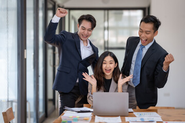 Asian business team celebrating success with raised fists at office desk with laptop and documents....