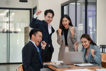 Asian business team celebrating success with raised fists at office desk with laptop and documents....