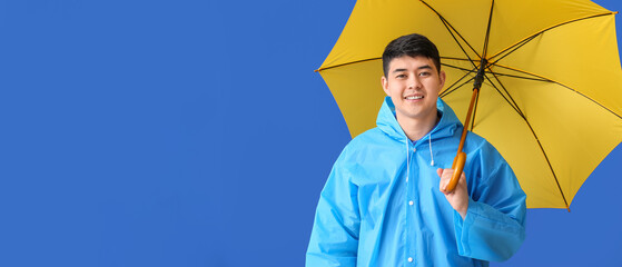 Young Asian man in raincoat and with umbrella on color background
