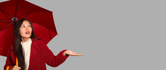 Thoughtful young Asian woman with umbrella on grey background