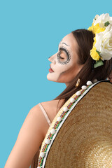 Young woman with painted skull on her face and sombrero hat against light blue background....