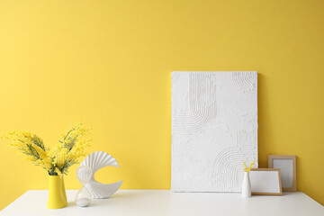 Vase with beautiful mimosa flowers, blank picture frames and decor on light table near yellow wall
