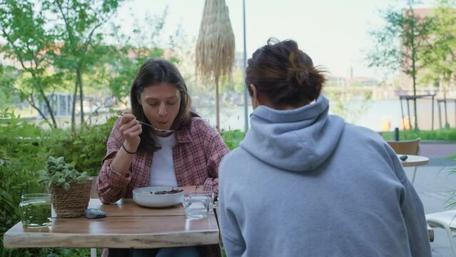 Quality time with friends. Two girls enjoying conversation, their healthy organic vegan acai bowl with fruits. Young woman sharing with her friend over lunch in hipster cafe. Female friendship concept
