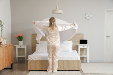 Pretty young woman in pajamas making bed in light bedroom