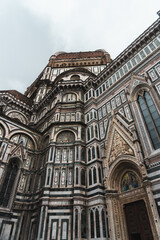 Santa Maria del Fiore Cathedral's ornate marble facade, blending Gothic and Renaissance styles, topped with its iconic dome. Stained glass windows and religious imagery adorn it under the cloudy sky.