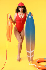 Beautiful young happy African-American female lifeguard with surfboard and ring buoy on yellow...
