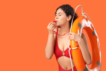 Beautiful young African-American female lifeguard with ring buoy whistling on orange background
