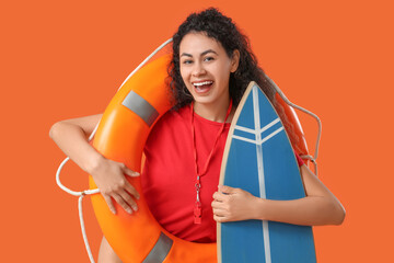 Beautiful young happy African-American female lifeguard with ring buoy and surfboard on orange...