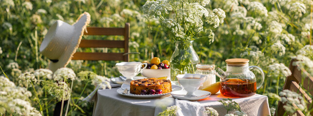 Elegant glamour table setting outdoor in the garden. White porcelain cups, teapot with herbal tea,...