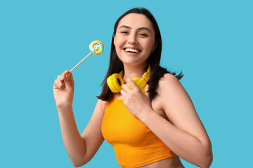 Happy young woman with sweet lollipop and headphones on blue background