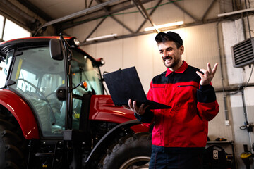 Experienced mechanic holding diagnostic laptop computer and servicing tractor agricultural machine.