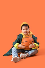 Cute little Asian boy in adult clothes with ball sitting on orange background