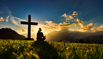 Silhouette of a young woman sitting on the grass praying in front of a cross at sunset and beautiful orange colored sky