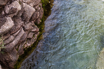 River flowing along a rocky cliff next to a waterfall, with moss and reflections in the water. View from above. Background. Texture. Horizontal.