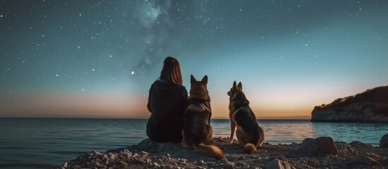 Woman and two german shepherd dogs stargazing on coastal beach at night, sitting together, rear view