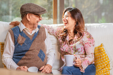 A beautiful young Latin woman enjoys a cup of coffee with her older father on the sofa in their...