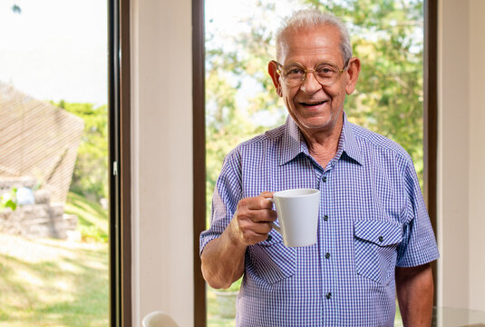 Portrait of an elderly man drinking coffee in front of a window where you can see the backyard of her house.