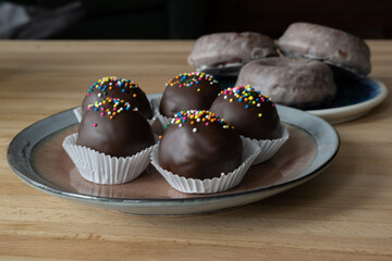 A small round plate of round double chocolate covered truffle balls with colorful sprinkles next to a blue plate with three chocolate glazed baked donuts. The sweets are on a wooden oak table. 