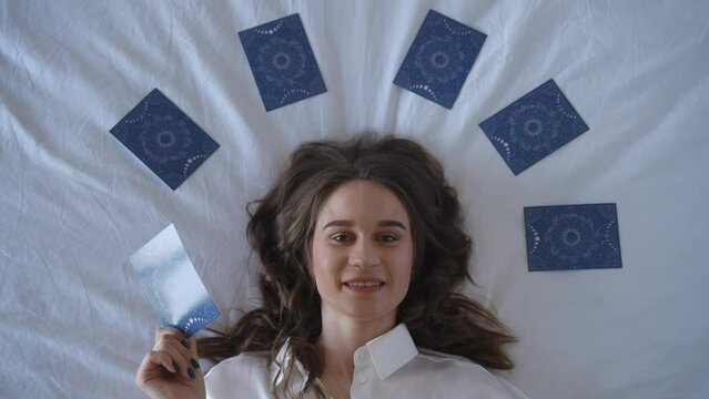 Top view. A woman in a white shirt lies on a white background surrounded by tarot cards and covers her face with the card she holds in her hand. The female astrologer removes her hand with the card