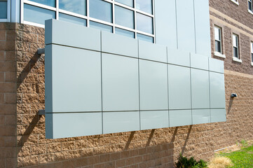 The exterior wall of a light green contemporary commercial style building with aluminum metal composite panels and small glass windows. The university dorm has a light brown brick exterior wall.