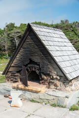 A vintage outdoor wood fired bread oven. The exterior structure is made from brick and rocks with a circular heavy metal door with an iron and wood handle.There's black soot on the hearth of the stove