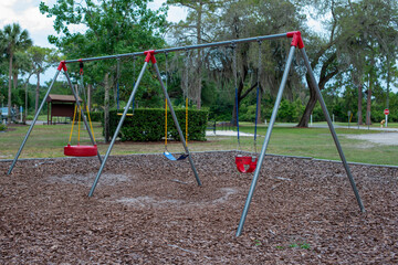 Multiple red plastic and rubber swings hanging from metal chains in a children's park. The background has large lush trees and benches. The ground is covered in tree mulch. 