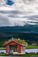A traditional quaint red bus shelter with a green sod roof stands before a mountainous dramatic cumulus clouds backdrop in the village Halsanaustan (Halsa), Trøndelag Norway