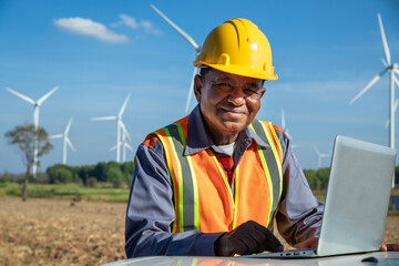 Male elderly engineer wearing a safety vest, uniform and yellow helmet using laptop in a wind farm...