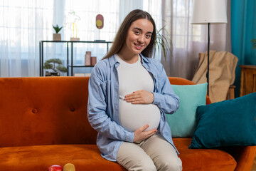 Portrait of smiling future mother sitting on couch touching pregnant belly in living room at home....