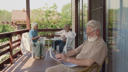 Medium full footage of elderly pensioners sitting outdoors on terrace in summer, Caucasian man in glasses, with grey beard reading newspaper in armchair, and couple chatting in background - Powered by Adobe