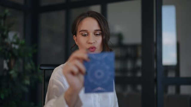 A female astrologer takes a tarot card from the table and reads what is written on the back while sitting at her desk in her office. The woman smiles while looking at the camera. Close-up