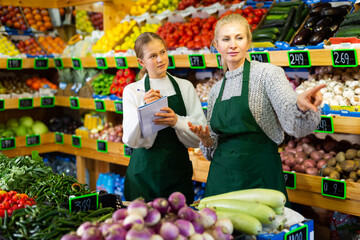 Saleswoman instructing her daughter before starting work in family store of fruits and vegetables,...