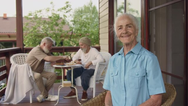 Medium portrait shot of senior Caucasian female resident relaxing in armchair on terrace at retirement home, knitting, looking at camera, smiling, male companions playing chess in background