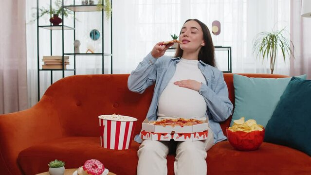 Pregnant woman eating unhealthy junk food pizza, popcorn crisp sitting on sofa watching TV movie in home living room. Fast fatty food during pregnancy in apartment. Hunger, change in taste preferences