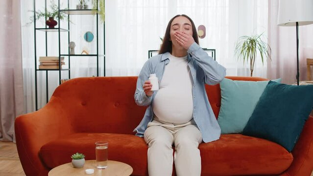 Pregnant Caucasian woman taking drug and drinking water to nourish unborn child sitting on sofa couch in living room at home. Health care medical and pregnancy concept. Lady happily looks at camera.
