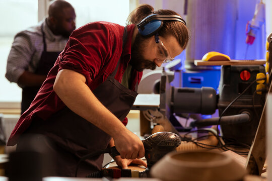 Artisan in joinery wearing safety earmuffs using orbital sander on lumber. Man in carpentry shop uses angle grinder to create wooden objects helped by colleague in blurry background