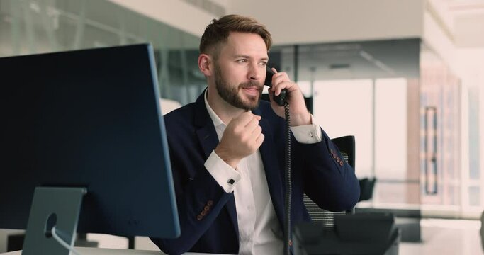 Millennial businessman, office employee sitting at workplace desk with PC holds telephone handset, answers incoming calls from company clients, provide professional help, information, support remotely