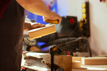 Craftsperson at workbench using manual sandpaper to sander lumber block, assembling furniture in...