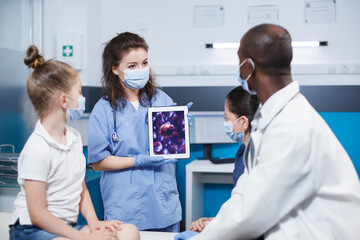 Woman wearing blue scrubs holding a digital tablet presenting a virus cell picture in front of a girl and her mother, and the african american male physician. Expert treatment and education provided. - Powered by Adobe