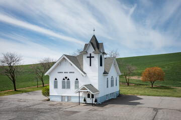 A small white church with a cross on top. The church is surrounded by trees and grass. The sky is blue and there are clouds in the background - 786754864