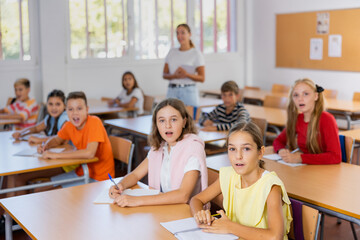 Enthusiastic surprised tween schoolchildren sitting at school desks at lesson, listening...