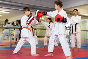 Kids in kimono, boxing gloves and focus mitts exercising jabs on group karate training. Their...