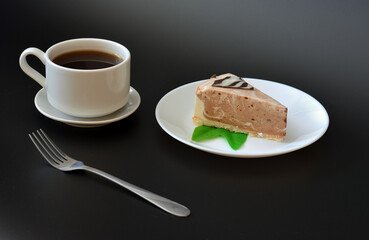 A piece of cream cheesecake with mint on a plate, a fork and a cup of hot black coffee on a saucer on a black background.
